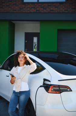 Evinde elektrikli arabasını şarj ederken akıllı telefonunu kullanan genç bir kadın sürdürülebilir ve çevre dostu ulaşımı kucaklıyor.