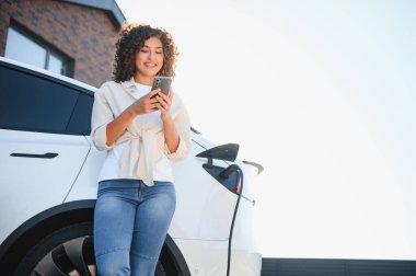 Elektrikli arabası evinin önünde şarj olurken cep telefonu kullanan mutlu kadın sürdürülebilir ve yeşil enerjinin tadını çıkarıyor.