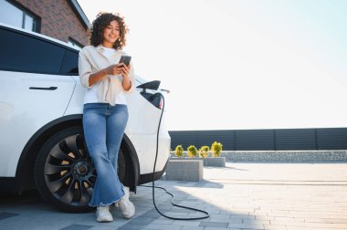 Modern evinin önünde elektrikli arabası şarj olurken akıllı telefon kullanan mutlu bir kadın.