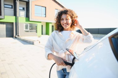 Gülümseyen kadın evinde şarj istasyonu kullanarak elektrikli aracını şarj ediyor, sürdürülebilir ulaşım ve yeşil enerji çözümlerini destekliyor