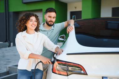 Genç çift modern evlerinde elektrikli araçlarını şarj ediyor, çevre dostu ulaşım ve yenilenebilir enerji çözümlerine bağlılıklarını sergiliyorlar.