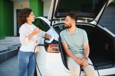 Çift evde elektrikli araçlarını şarj ederken sohbet ediyor, sürdürülebilir ulaşım ve yenilenebilir enerjiyi kucaklıyor