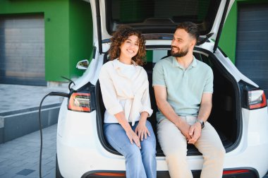 Genç çift elektrikli arabalarının açık bagajında oturuyor, gülümsüyor ve evde şarj olmanın keyfini çıkarıyorlar.