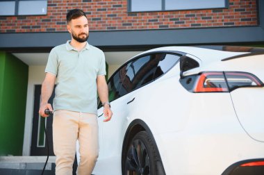 Gururlu ev sahibi, modern evinin önüne park ettiği Tesla Model Y elektrikli arabasından elektrik kablosunu kesiyor.