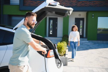 Kadın sürdürülebilir yaşam tarzı ve yeşil enerji sergilerken, adam modern evin önünde elektrikli arabaya elektrik kablosu takıyor
