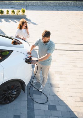 Kadın telefonda konuşurken, adam elektrik kablosunu elektrikli araca bağlıyor, sürdürülebilir yaşam tarzı ve çevre dostu ulaşım gösteriyor