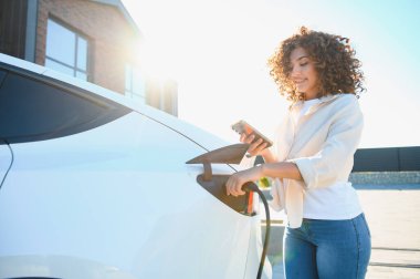 Gülümseyen genç kadın elektrikli arabasını evde şarj ediyor, sürdürülebilir ve yenilenebilir enerjiyi çevre dostu yaşam için kucaklıyor
