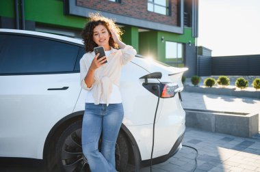Genç bir kadın elektrikli arabası modern evinin önünde şarj olurken akıllı telefonunu kullanıyor.