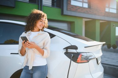 Genç bir kadın elektrikli arabasını şarj ederken cep telefonu uygulaması kullanıyor. Kullanışlı ve sürdürülebilir ulaşımı gösteriyor.