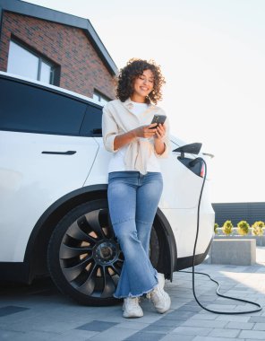 Modern bir evin önünde elektrikli arabasını şarj ederken akıllı telefon kullanan genç bir kadın yenilenebilir enerji ve sürdürülebilir yaşam enerjisini ön plana çıkarıyor.