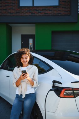 Elektrikli arabası modern evinin önünde şarj olurken akıllı telefon kullanan mutlu kadın sürdürülebilir ve yeşil enerjinin tadını çıkarıyor.