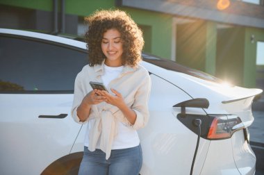 Elektrikli arabası evde şarj olurken akıllı telefonunu kullanan genç bir kadın sürdürülebilir ve kullanışlı ulaşımın keyfini çıkarıyor.