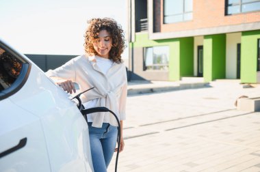 Genç kadın elektrik kablosunu elektrik aracına bağlıyor, çevre dostu ulaşımı ve yenilenebilir enerji çözümlerini teşvik ediyor