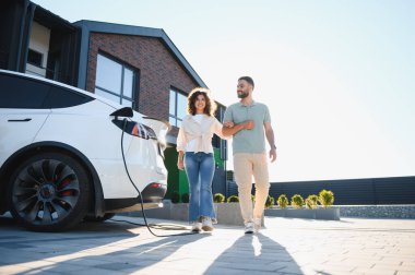 Genç bir çift elektrikli arabalarının yanında yürüyor ve el ele tutuşuyor. Modern evlerinin önüne doluşuyor, sürdürülebilir ve çevre dostu yaşam tarzlarını kucaklıyor.