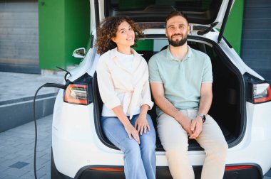 Elektrikli arabanın açık bagajında oturan mutlu çift, evlerinin yakınında akünün şarj olmasını bekliyor, sürdürülebilir yaşam tarzını kucaklıyorlar.