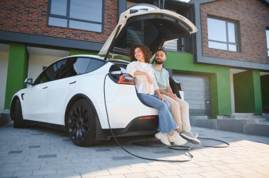 Elektrikli arabalarının açık bagajında oturan mutlu çift, modern evlerinde şarj oluyorlar, sürdürülebilir yaşam tarzının tadını çıkarıyorlar.