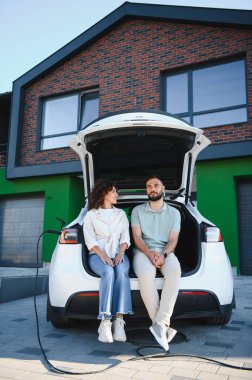 Genç bir çift elektrikli arabalarının bagajında oturuyor, modern evlerinde şarj oluyorlar, sürdürülebilir ulaşımın tadını çıkarıyorlar.