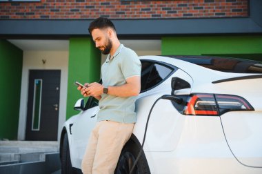 Adam elektrikli arabaya yaslanıyor, şarj olurken akıllı telefonu kontrol ediyor.