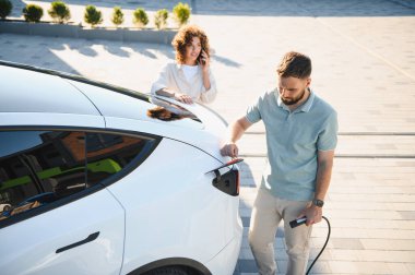 Kadın arka planda telefonla sohbet ederken, adam elektrik kablosunu elektrik kablosuna takıyor ve modern sürdürülebilir yaşamı ön plana çıkarıyor.