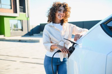 Genç kadın elektrik kablosunu elektrik aracına bağlıyor, eko bilinçli ulaşım ve yenilenebilir enerji çözümlerine geçişi vurguluyor