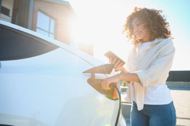 Gülümseyen kadın elektrikli aracını şarj istasyonunda şarj ediyor, yenilenebilir enerji ve sürdürülebilir yaşam enerjisini kucaklıyor