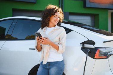 Evinin yakınında elektrikli araba şarj ederken akıllı telefon kullanan gülümseyen genç bir kadın. Yenilenebilir enerjiyi ve sürdürülebilir yaşamı kucaklamak