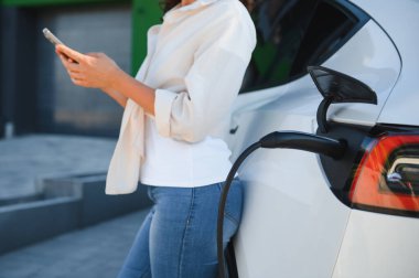 Genç bir kadın elektrikli arabasının yanında duruyor, istasyonda şarj ediyor, yenilenebilir enerjiyi ve sürdürülebilir yaşamı simgeliyor.