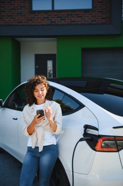 Genç bir kadın modern evinde elektrikli arabasını şarj ederken akıllı telefon uygulaması kullanıyor, sürdürülebilir ulaşımı kucaklıyor.