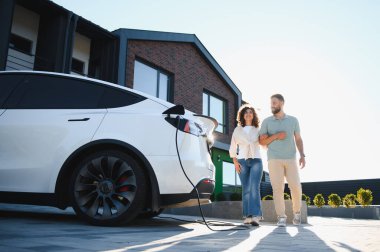 Elektrikli arabaları dışarıda şarj olurken mutlu çift modern evlerinin yanında yürüyor.