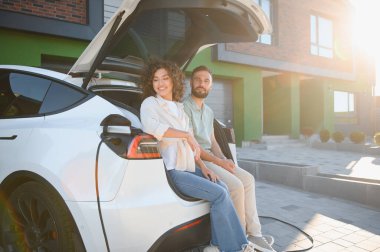 Elektrikli arabalarının açık bagajında oturan mutlu çift, modern evlerinde şarj oluyorlar, sürdürülebilir ve çevre dostu yaşam tarzlarının tadını çıkarıyorlar.