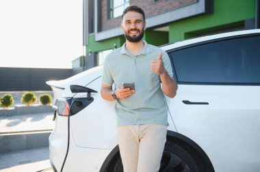 Akıllı telefonu tutan ve evde elektrikli arabayı şarj ederken baş parmağını gösteren bir adam.