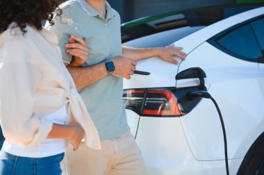 Evlerinde şarj olan elektrikli arabalarının yanında duran çift sürdürülebilir ulaşım ve yenilenebilir enerjiye olan bağlılıklarını gösteriyor.