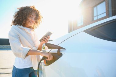 Modern evin dışında elektrikli araba şarj ederken akıllı telefon kullanan gülümseyen kadın, yenilenebilir enerji ve sürdürülebilir yaşamı kucaklıyor