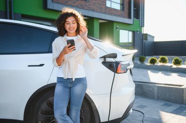 Modern evin önünde elektrikli araba şarj ederken akıllı telefon kullanan genç kadın, sürdürülebilir ulaşım ve yeşil enerjiyi kucaklıyor