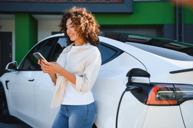Elektrikli arabası evinin önünde şarj olurken akıllı telefon kullanan genç bir kadın, sürdürülebilir ulaşım ve yeşil enerji çözümlerini kucaklıyor