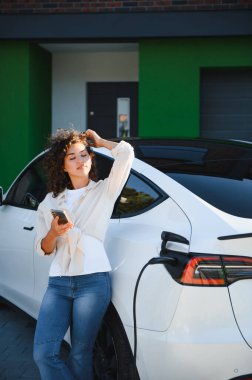 Genç bir kadın elektrikli arabasını şarj ederken akıllı telefon kullanıyor. Sürdürülebilir ve kullanışlı ulaşımı gösteriyor.