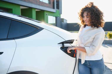 Gülümseyen genç kadın elektrikli aracını şarj istasyonunda şarj ediyor, yenilenebilir enerji ve sürdürülebilir yaşam enerjisini vurguluyor