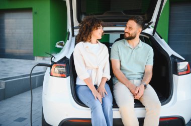 Elektrikli arabanın açık bagajında oturan mutlu çift, arabalarının şarj olmasını beklerken sohbetin tadını çıkarıyorlar, sürdürülebilir yaşam tarzlarını kucaklıyorlar.