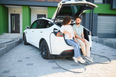 Genç çift elektrikli arabalarının açık bagajında oturmuş, evlerinde şarj oluyorlar, sürdürülebilir ulaşımı kucaklarken birlikte bir anın tadını çıkarıyorlar.