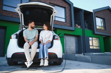 Elektrikli arabalarının açık bagajında oturan mutlu çift, modern evlerinde şarj oluyorlar, sürdürülebilir ve çevre dostu yaşam tarzlarının tadını çıkarıyorlar.