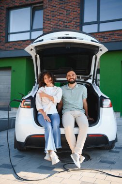 Genç çift açık bagajda dinlenirken elektrikli arabaları modern evlerinin yanında şarj oluyor ve sürdürülebilir ulaşımı kucaklıyor.
