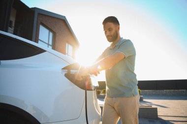Modern bir evin dışındaki şarj aletine elektrikli araba bağlayan adam.