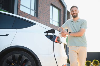 Evde şarj ederken elektrikli arabasına yaslanan bir adam.