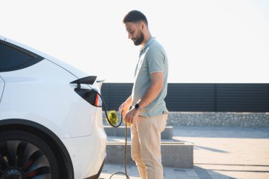 Açık hava istasyonundaki Tesla Model Y aracına şarj kablosu bağlayan sürücü