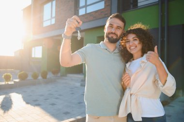 Genç ev sahipleri yeni modern evlerinin yanında dikilirken anahtarlarını gösterip başparmak işareti yapıyorlar.