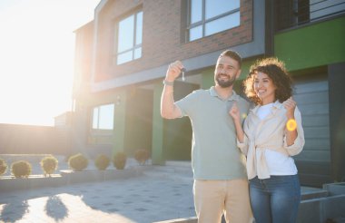 Neşeli ev sahipleri yeni evlerinin anahtarlarını gösteriyor, güneşli bir günde gayrimenkul alımını kutluyorlar.