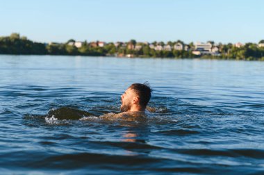 Gölde çırpınan sakallı bir adam, soluk soluğa kalan şehir silüeti arka planda belirdiğinde, çaresizlik ve tehlike anlarını yakalıyor.