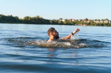 Sakallı bir adam gölde ayakta kalmaya çalışıyor, nefes nefese ve yanlış giden yaz tatili boyunca panikle boğuşuyor.