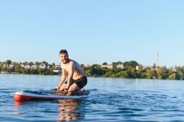 Genç adam, dalgalı ağaçlar ve açık mavi gökyüzü ile çevrili sakin bir gölde kürek çekerken güneşi emerek kürek çekiyor.
