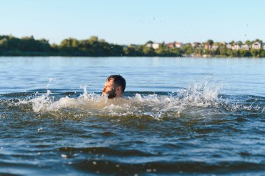 Sakallı bir adam yaz günü nehir suyunda boğuşuyor, nefes nefese kalıyor ve akıntıya karşı savaşırken su sıçratıyor.